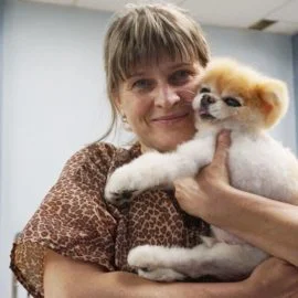 A woman with light brown hair smiles while holding a small, fluffy dog with a trimmed coat in her arms indoors, showcasing the results of a top-notch pet groomer.