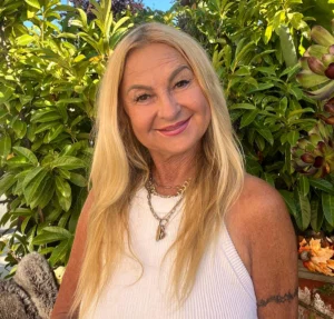 Smiling woman with long blonde hair in a white top, standing outside in front of green plants and blue sky, radiates confidence—just like ourGroomers' happy clients.