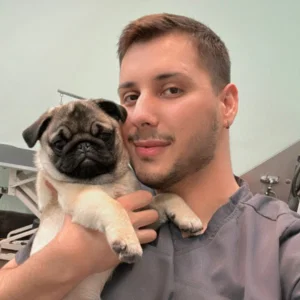 A man in scrubs, one of ourGroomers, holds a small pug puppy and smiles for a selfie in a veterinary clinic.